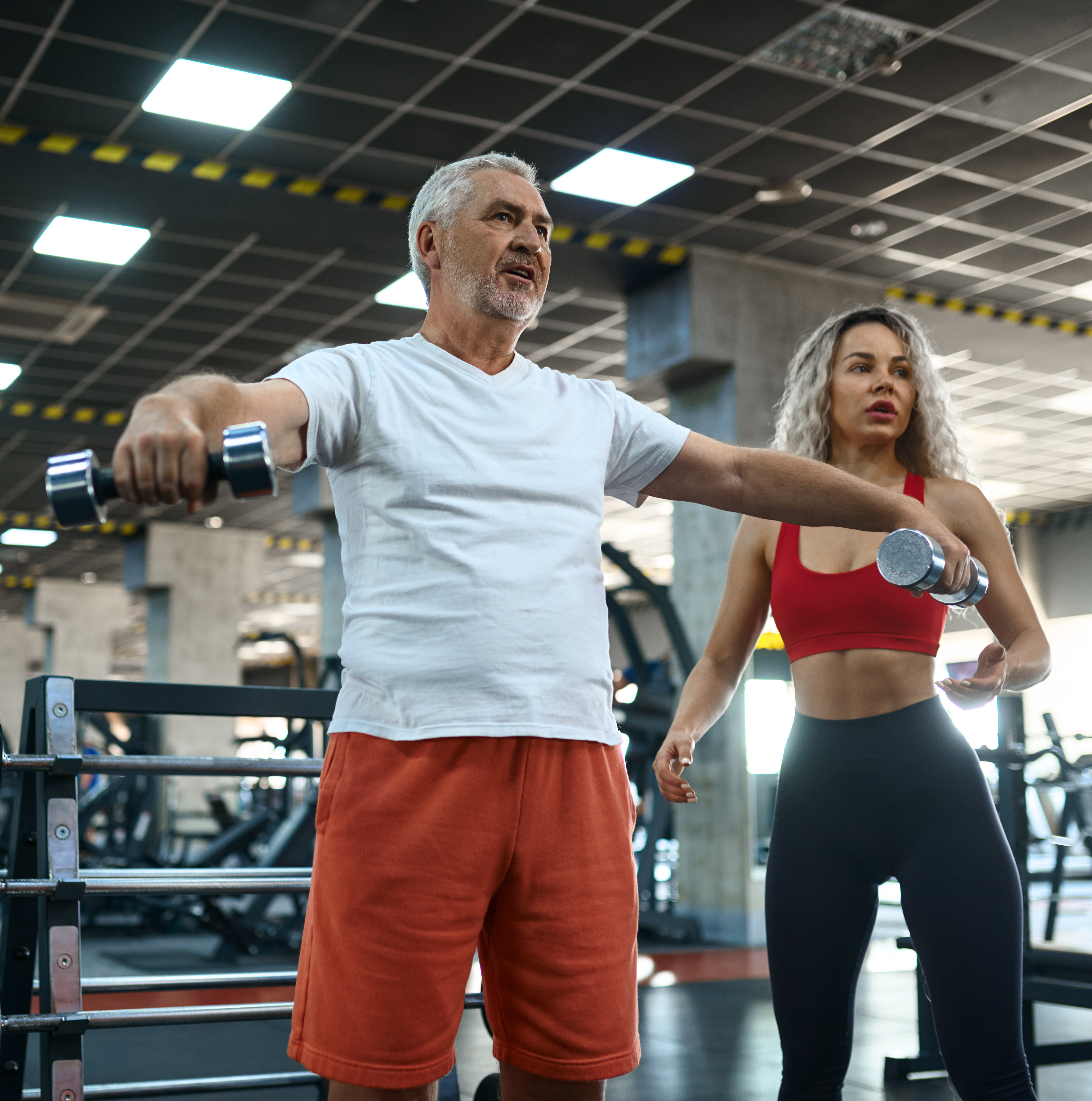 Old man doing exercise with dumbbells, female personal trainer, gym interior on background. Sportive grandpa with woman instructor, training in sport center